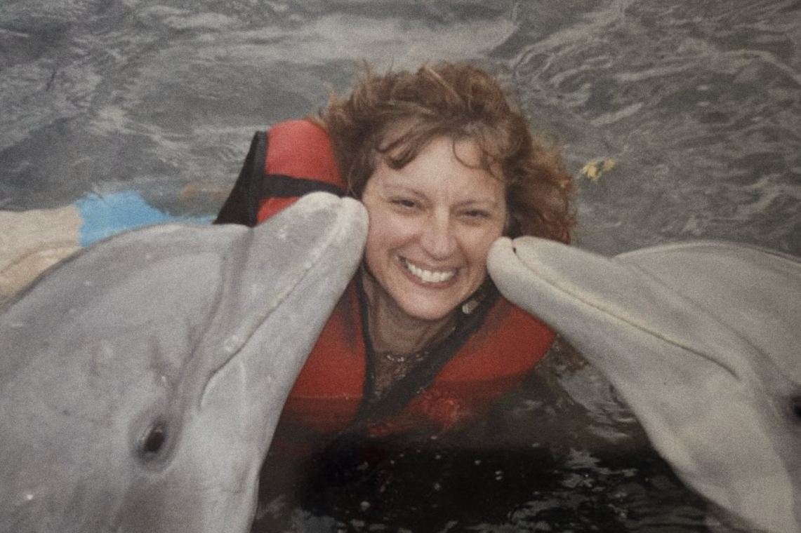 Josée en veste de sauvetage rouge dans l'eau, embrassée par deux dauphins.